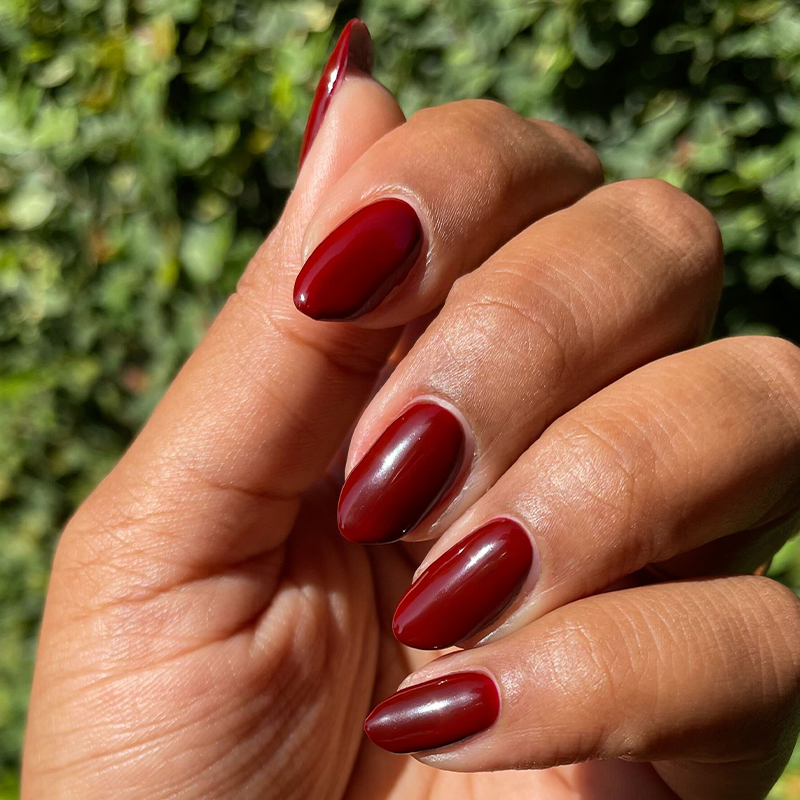 Hand with medium-length nails painted in glossy deep red polish from Chocolate Cherry manicure by Olive and June outdoors