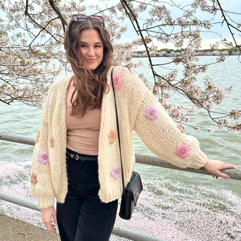 Woman wearing beige floral knit cardigan and black pants standing by waterfront in springtime with Ally Rooker’s Nail Essentials Bundle