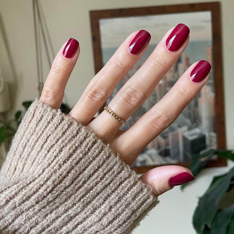 Hand with shiny mulberry gel polish manicure in rich berry shade wearing cozy knitted sweater indoors