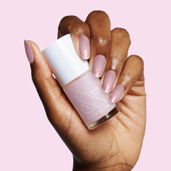 Hand with softly polished nails holding The Glazed Mani System nail polish bottle by Olive and June against pale pink background