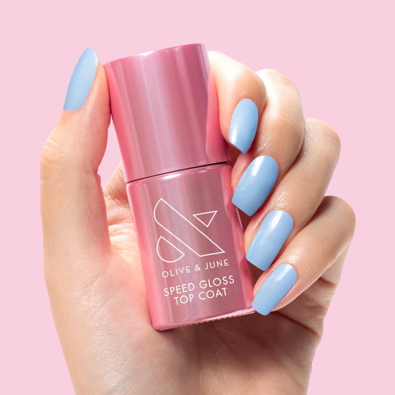 Hand holding a pink bottle of Speed Gloss Top Coat with glossy blue manicured nails on a pink background