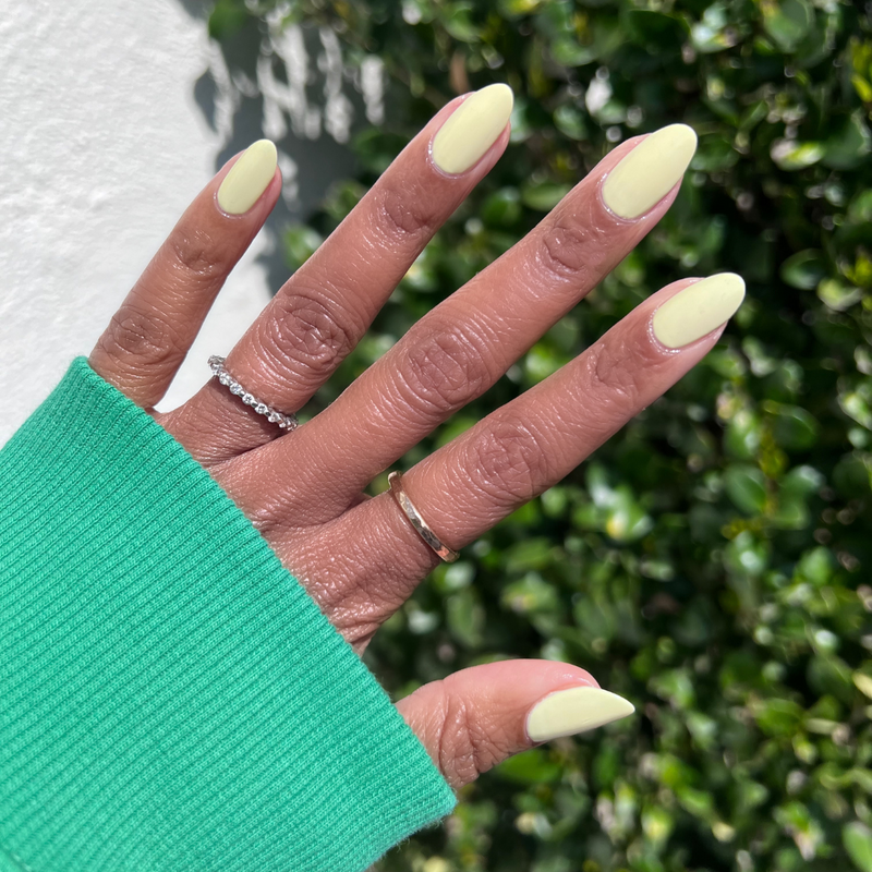 Hand with almond-shaped nails painted in Sunny pastel yellow polish showing a smooth, bright manicure outdoors with green foliage background