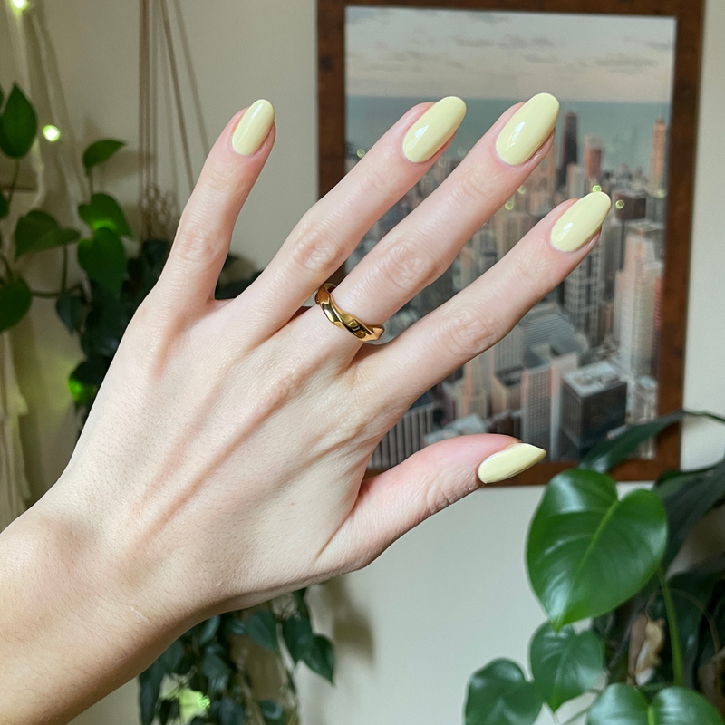 Hand with rounded nails painted in Sunny pastel yellow showing a smooth, bright manicure with indoor plants and city view background