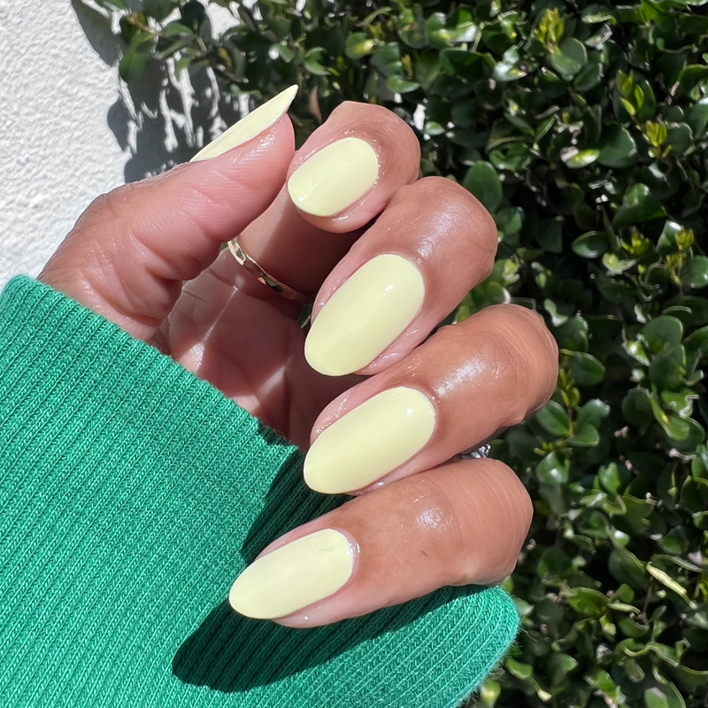 Hand with oval nails painted in Sunny pastel yellow polish showing a smooth, bright manicure outdoors with green foliage background