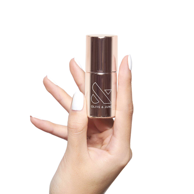 Hand holding Nail Primer bottle by Olive and June with white polished nails on white background
