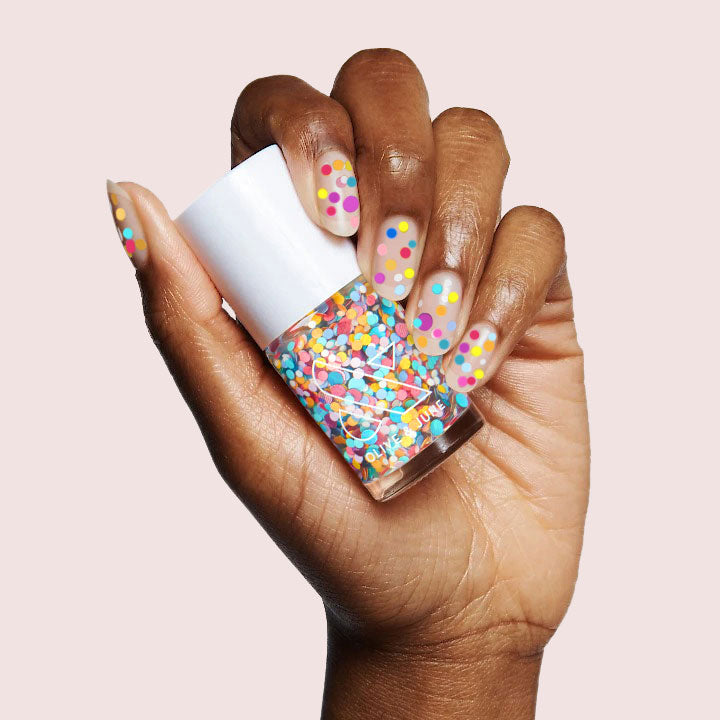 Hand with nails decorated in colorful dots holding Olive and June Sprinkle Cookie nail polish bottle on light background