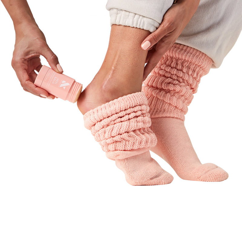 Person applying Heel Balm stick to dry heel while wearing cozy pink knit socks on white background