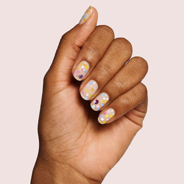 Hand with nails decorated in multicolored dots using Sprinkle Cookie polish on pale pink background for manicure