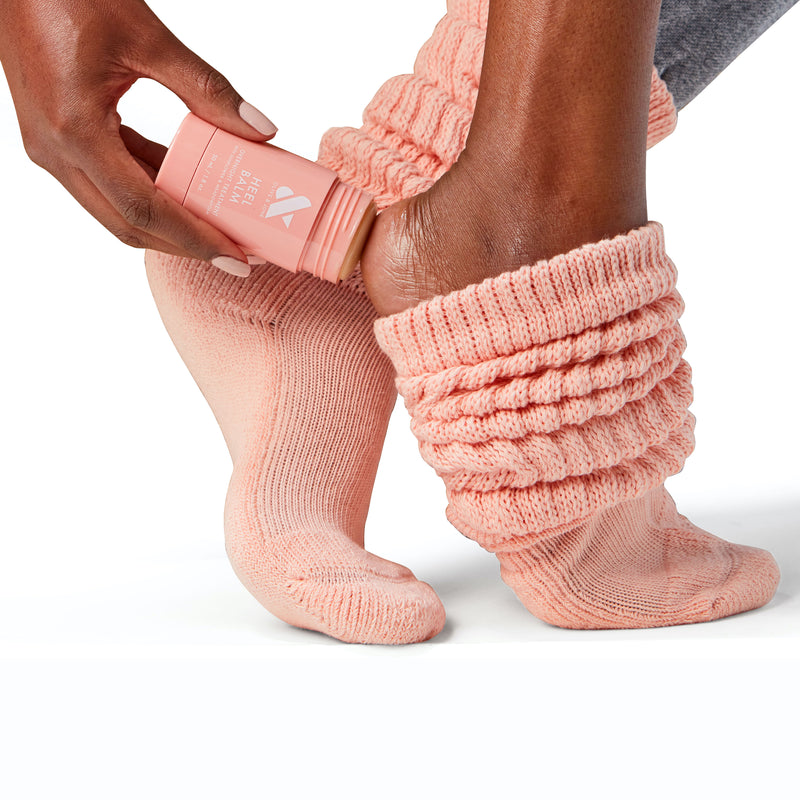Person wearing peach knit socks applying Heel Balm stick to heel for dry and cracked skin care treatment