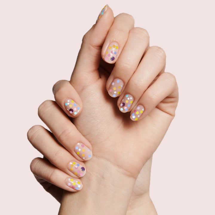 Hands with nails decorated in colorful confetti dots using Sprinkle Cookie polish on soft pink background for manicure