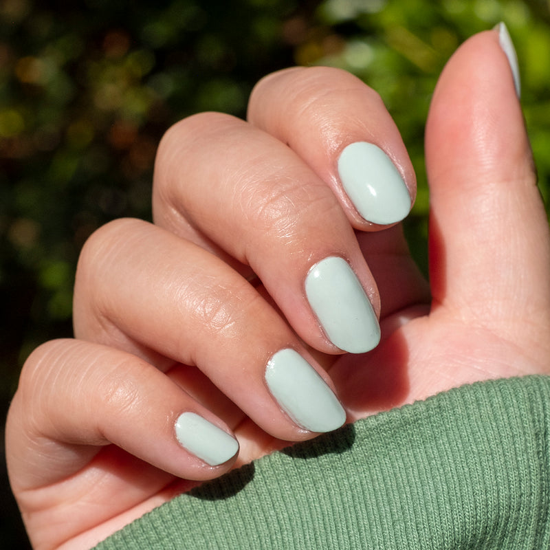 Hand with nails painted in soft sage green KMC gel polish showing a smooth, natural manicure in bright sunlight