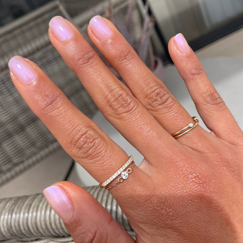 Hand with short nails coated in Baroque gel polish showing soft pearly iridescence with delicate gold rings on wicker background