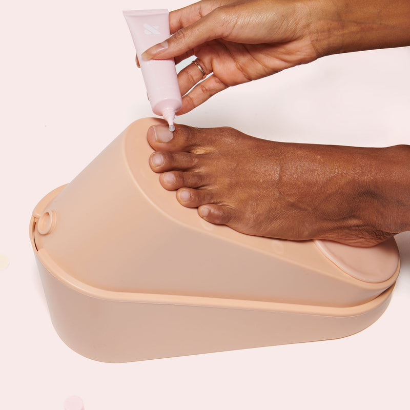 Hand applying Cuticle Remover from soft pink tube onto toenail on beige footrest for gentle nail care and mani prep