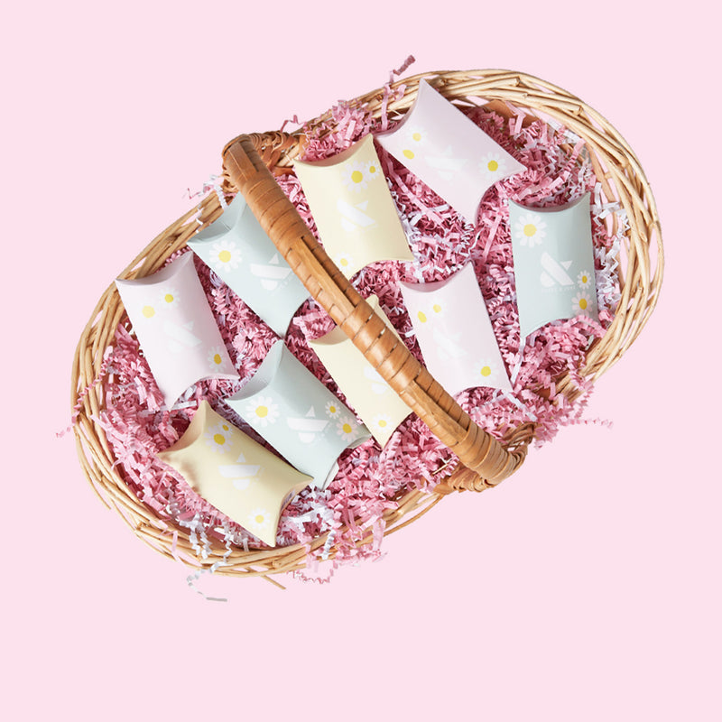 Wicker basket filled with pastel printed pillow boxes of the Spring Minis Set on pink shredded paper background
