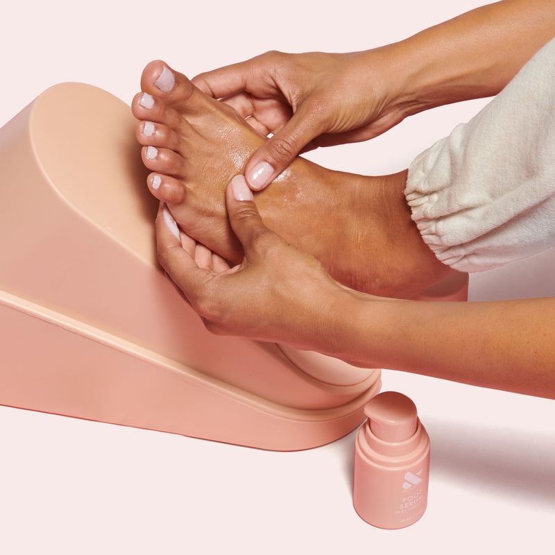 Person massaging foot with Foot Serum applied, seated on light pink footrest with soft pink serum bottle nearby