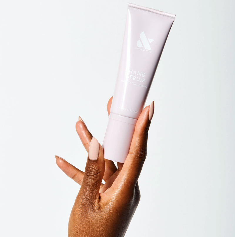 Hand holding a pale pink tube of Hand Serum by Olive and June with manicured nails in a neutral shade