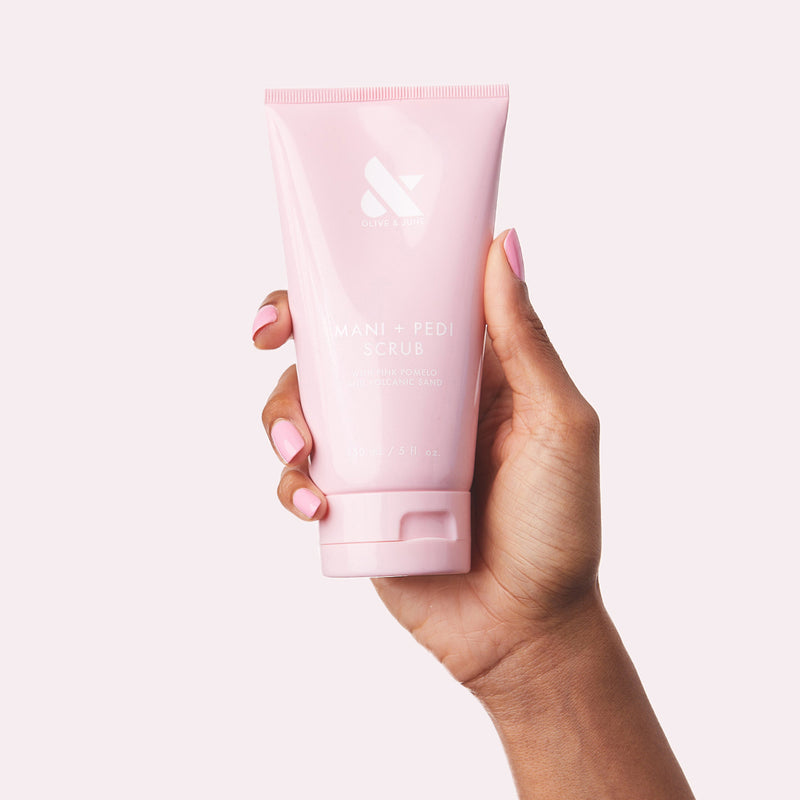 Hand holding pink tube of MANI + PEDI SCRUB with neatly manicured nails in soft pink polish against light background