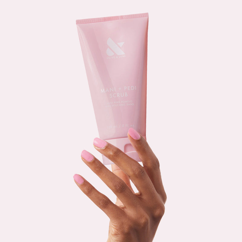 Hand with soft pink manicure holding pink tube of Olive and June MANI + PEDI SCRUB for nail care and exfoliation