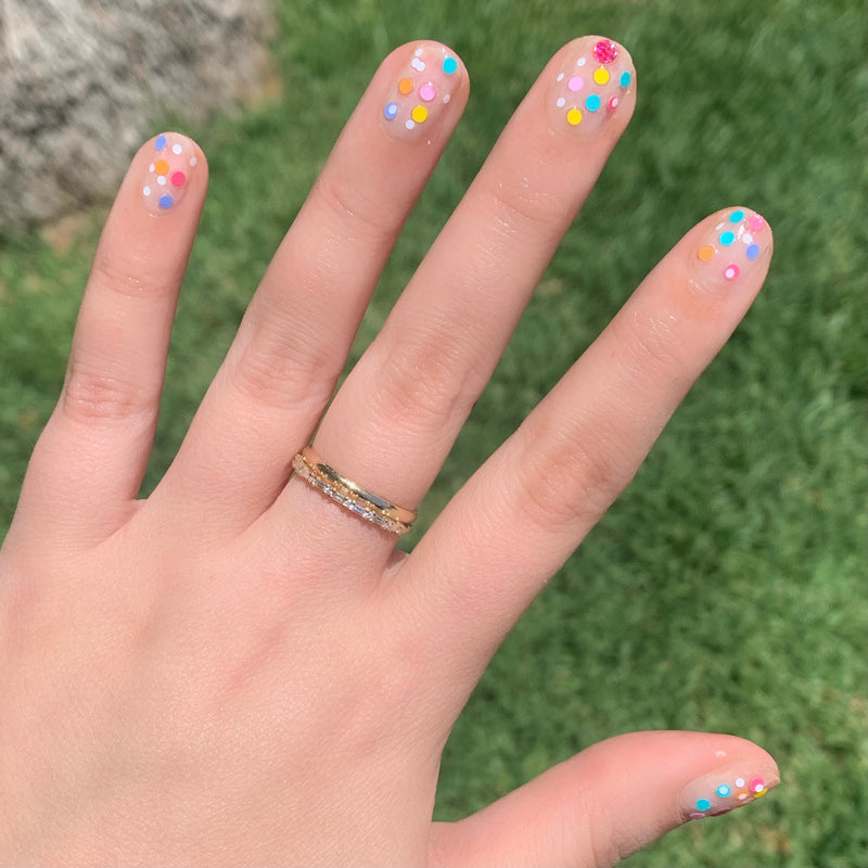 Hand with nails featuring colorful dots manicure using Sprinkle Cookie polish against outdoor green grass background