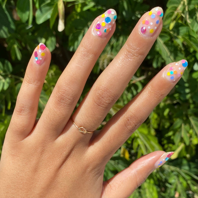 Hand with nails painted in Olive and June Sprinkle Cookie polish featuring colorful confetti dots against green leaves background