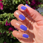 Hand with nails polished in vibrant royal purple House of O&J nail polish against green leaves and magenta flowers background