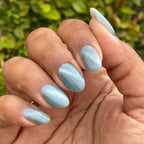 Fingernails painted with Aqua Velvet - Gel bright pastel blue magnetic gel polish showing shimmering velvet finish against natural greenery background