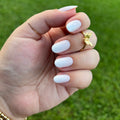Hand with oval-shaped nails painted glossy pale white held against blurred green grass background thumbnail
