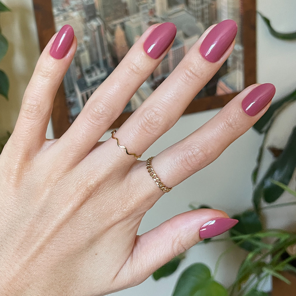 LD - Press-On | Medium | Oval showing hand with glossy medium oval rosy press-on manicure indoors with gold rings and plants background