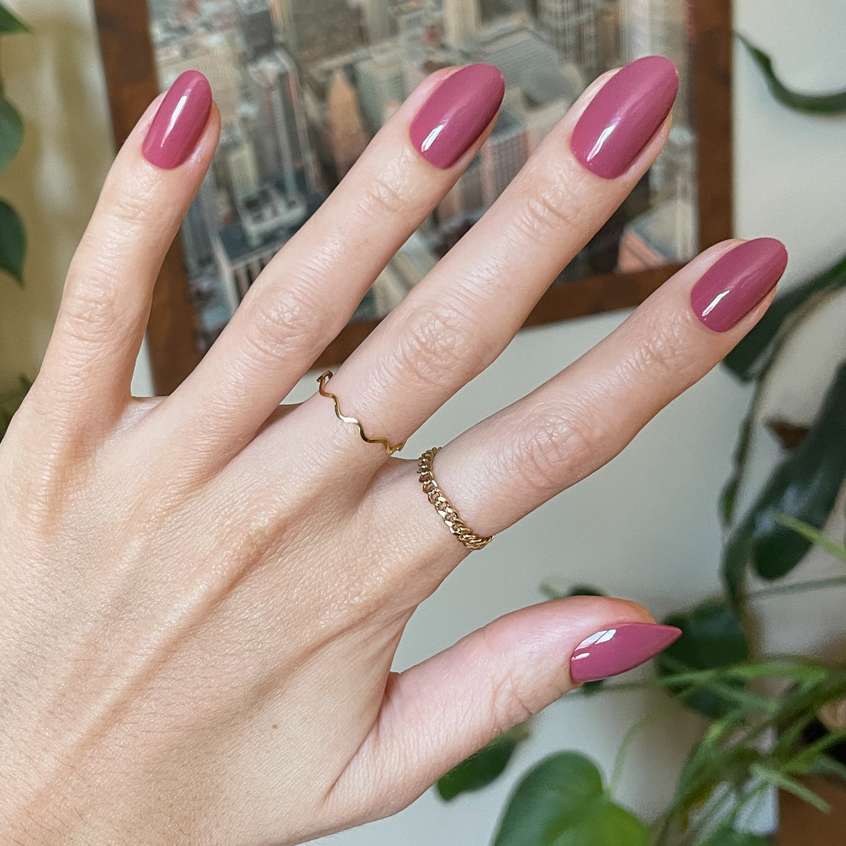 LD - Press-On | Medium | Oval showing hand with glossy medium oval rosy press-on manicure indoors with gold rings and plants background