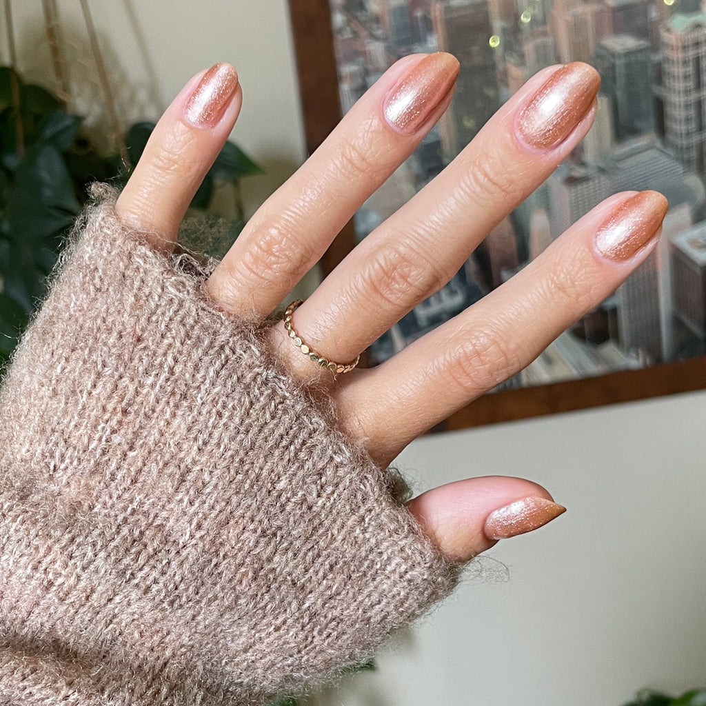 Hand with Penny polish showing a glossy copper metallic manicure on light skin wearing a fuzzy beige sweater sleeve