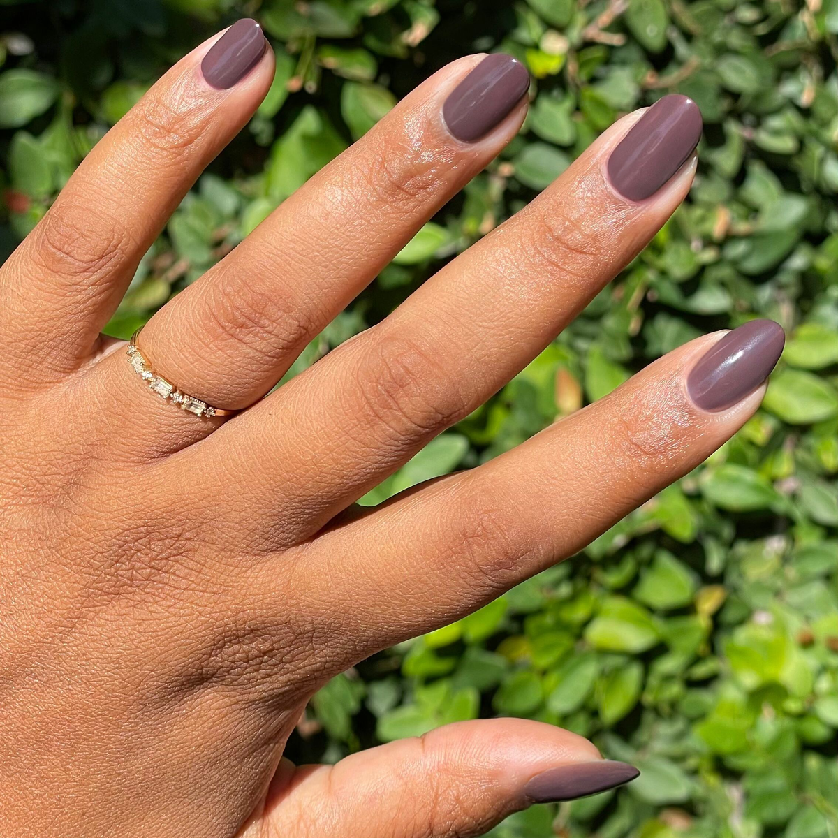 Bunny deep neutral brown gel polish on natural nails outdoors with greenery background and subtle gold ring