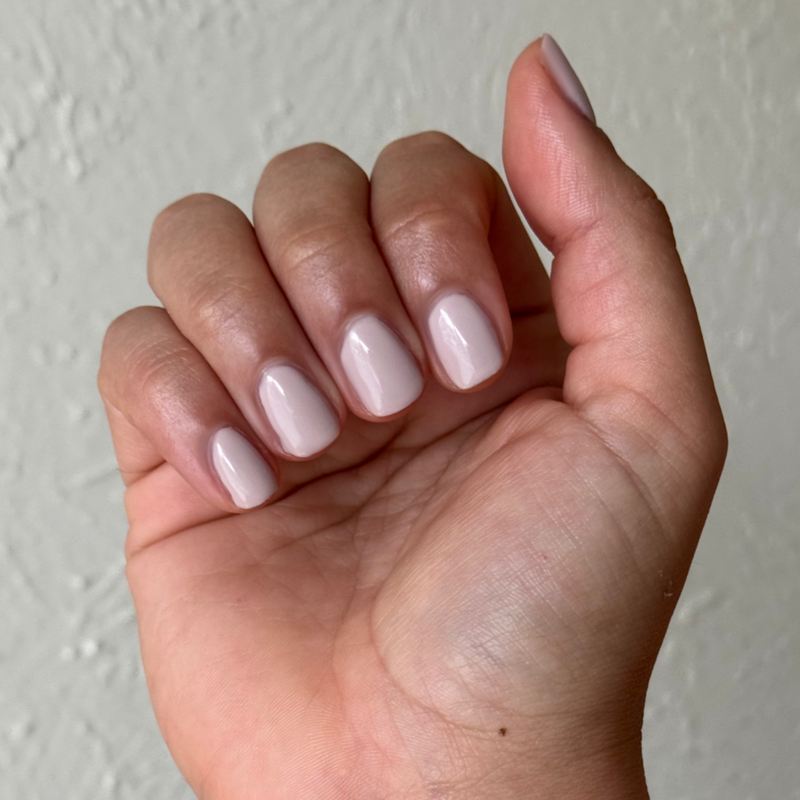 Hand with short rounded nails polished in CDJ - Gel creamy neutral gel polish against textured wall background