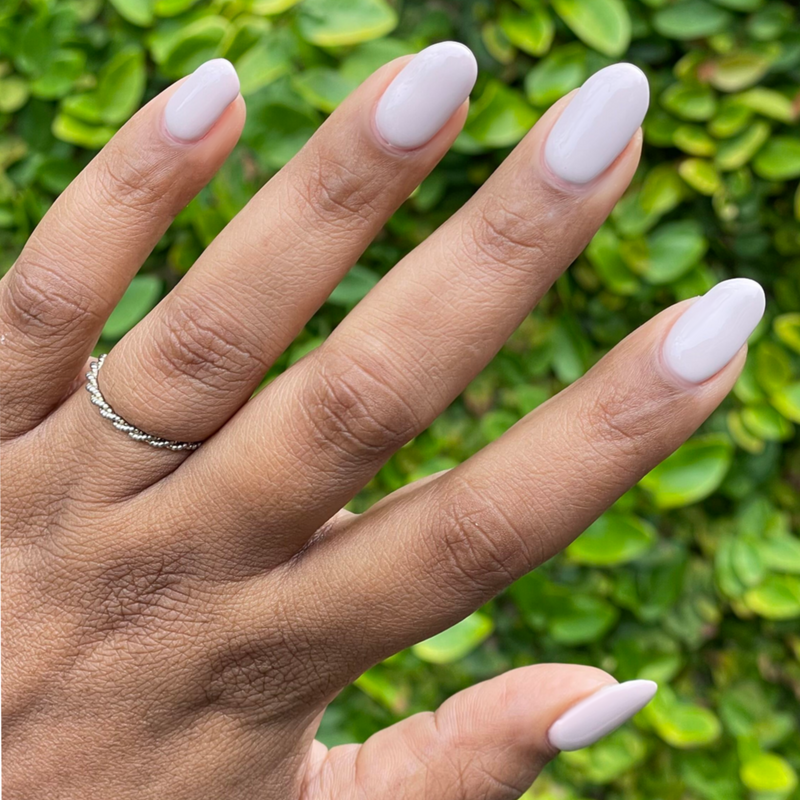 Hand with medium-length oval nails painted in CDJ - Gel creamy neutral gel polish against green leafy background