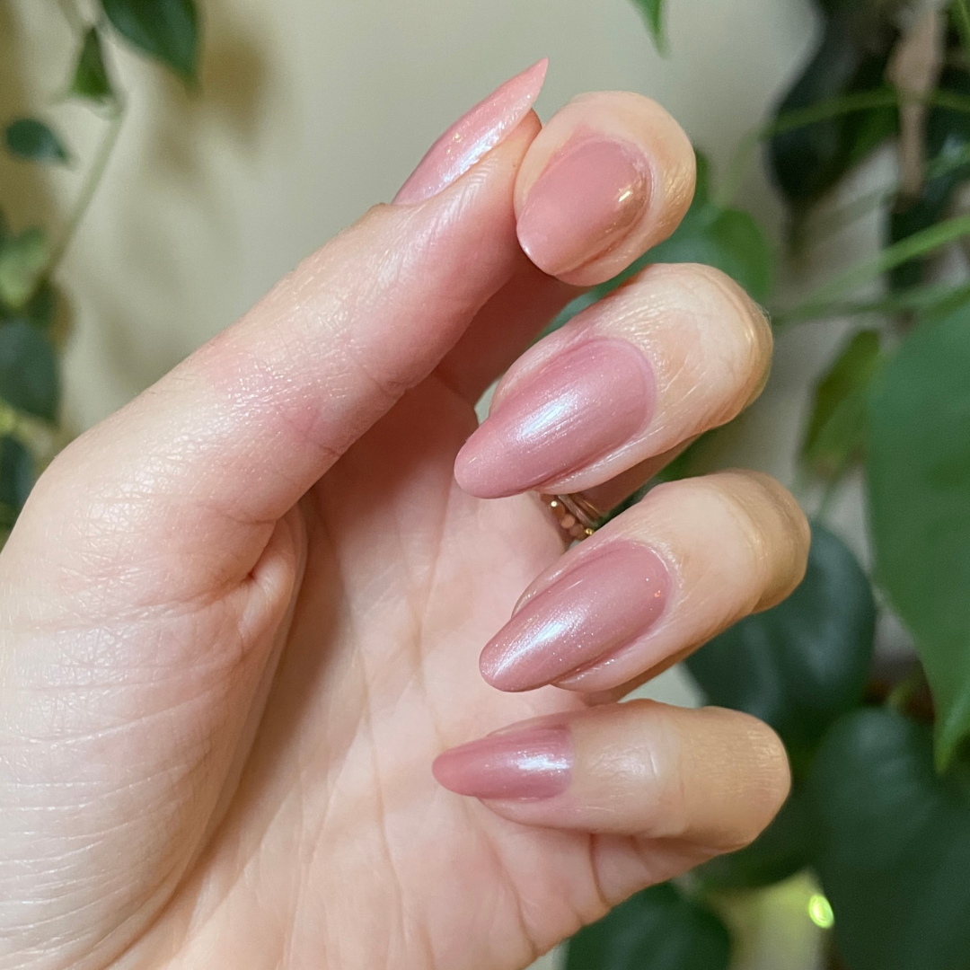 Hand with almond-shaped nails painted in Lovenote frosty pink pearl quick dry polish against blurred greenery background