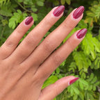 Hand with glossy Cabernet gel polish in rich plummy red showing a polished manicure against green foliage background