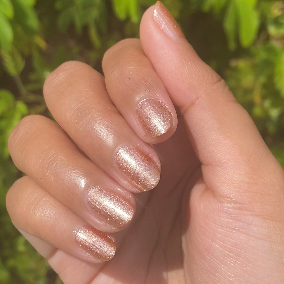 Hand with Penny showing a shiny copper metallic manicure on medium skin with natural outdoor lighting and green foliage background
