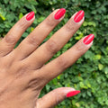 Hand with smooth red nails polished using Domino’s Pizza Mani Kit shown against green leafy background thumbnail