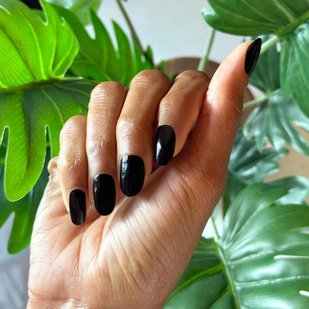 Hand with glossy Covet gel polish manicure in rich dark plum on medium-length nails against green leafy background