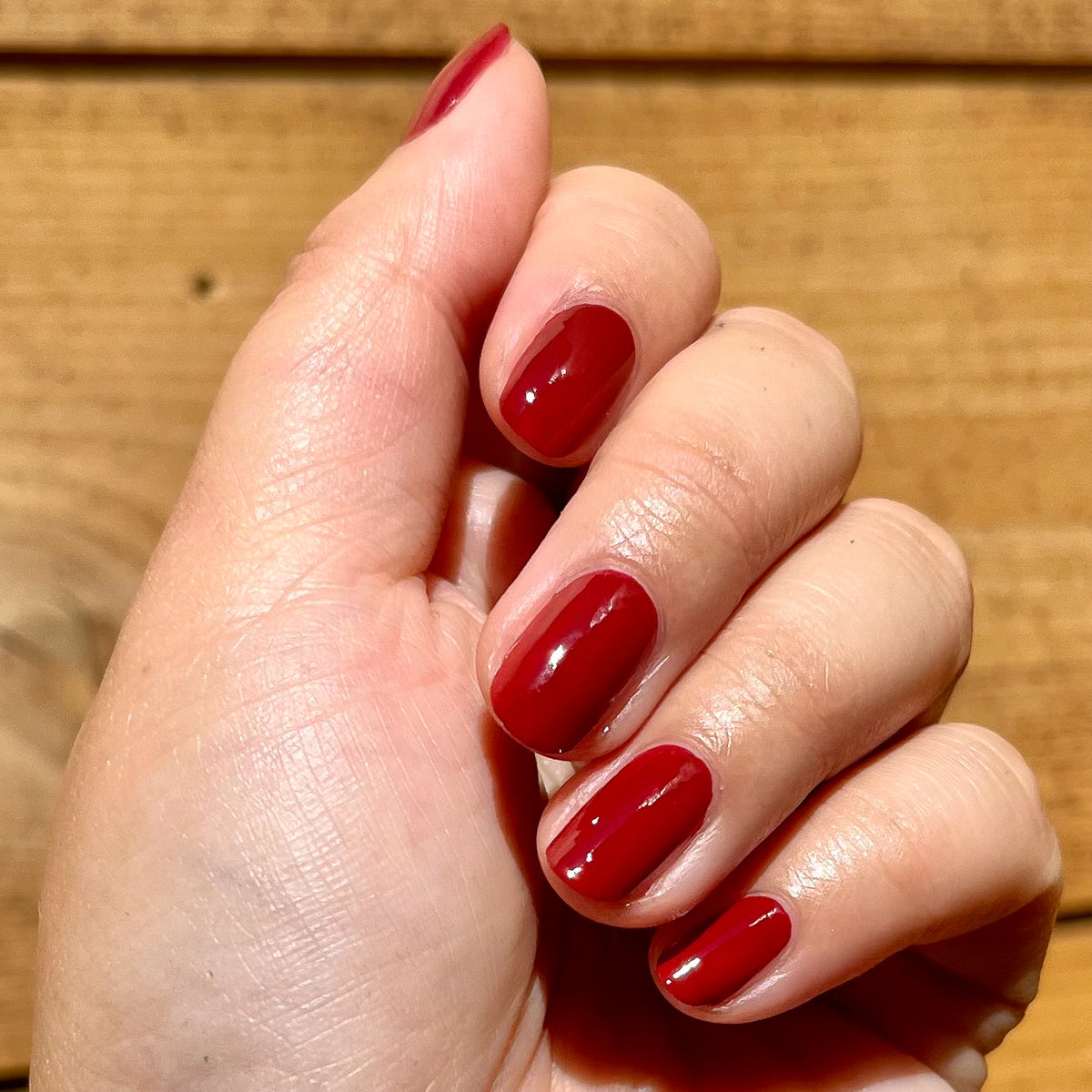 Hand showing glossy deep earthy red manicure with Brick polish on natural skin against a wooden background