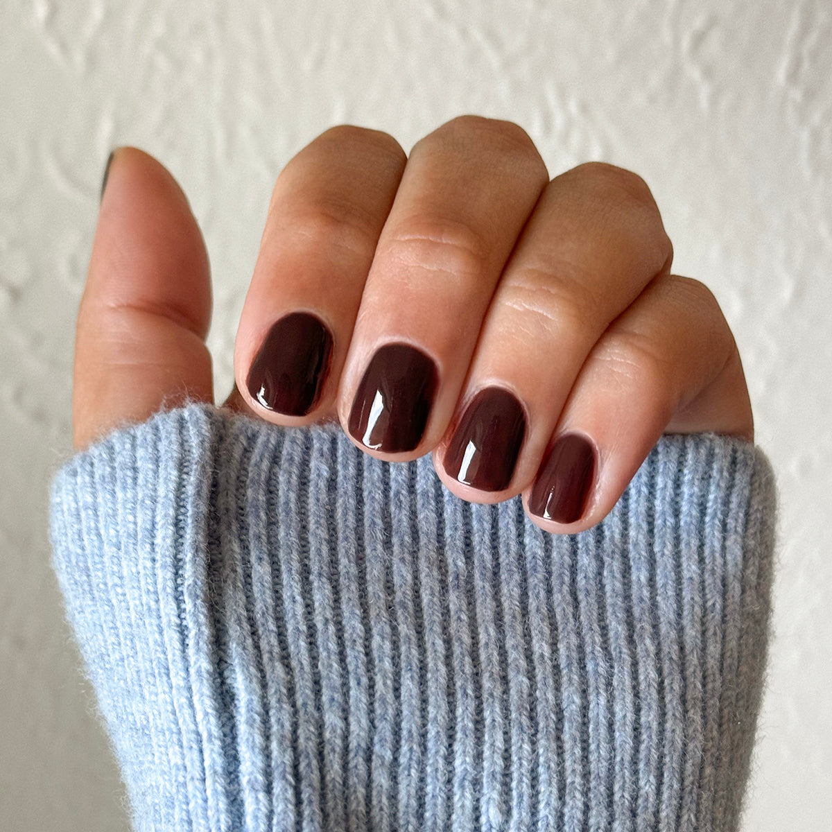 Hand with Dark Chocolate gel polish on short rounded nails against soft blue sweater background for cozy manicure look