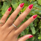 Hand with almond-shaped nails painted glossy Garnet Glass gel polish wearing gold rings against outdoor green foliage background