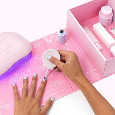 Hand applying light purple gel polish on nails using a brush with manicure kit and UV lamp on pink background
