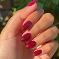 Hand with glossy deep red gel polish nails held against a blurred green outdoor background thumbnail