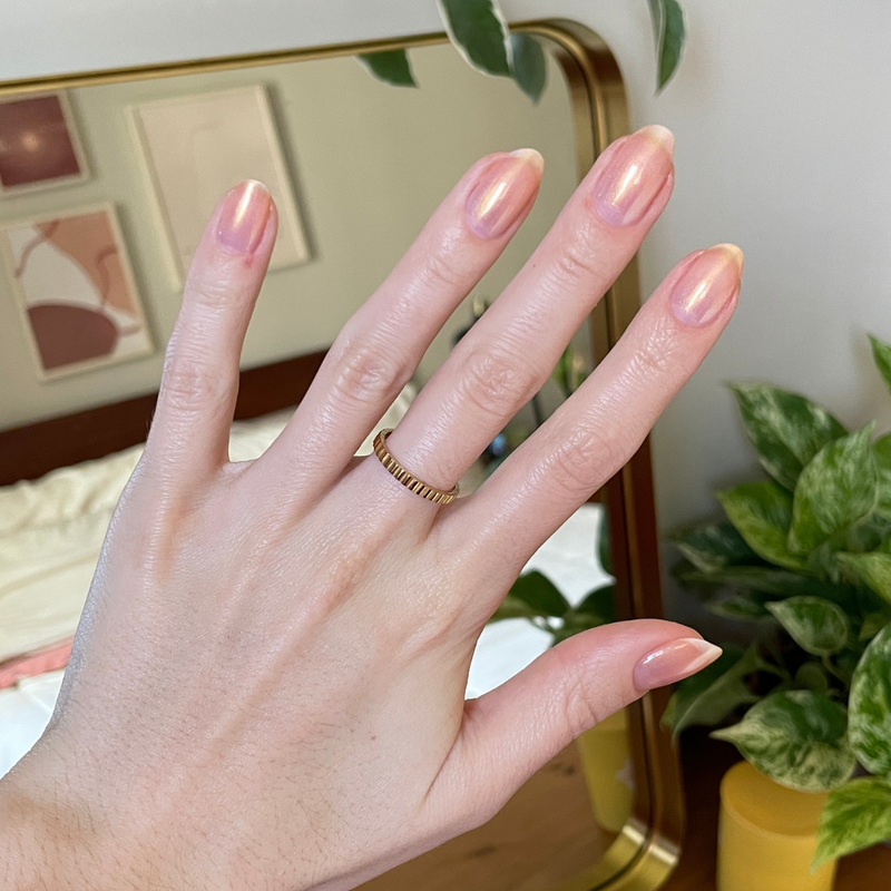 Hand with natural nails featuring a subtle shimmer from Gold Drops polish in front of a mirror and green plants for mani display