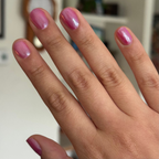 Hand with short nails painted in HARD LAUNCH pinky-purple iridescent duochrome polish on blurred indoor background
