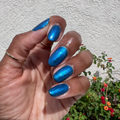 Hand with shiny blue nails featuring Emerald Chrome gel polish manicure in natural sunlight outdoors with plants background thumbnail