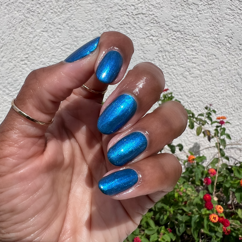 Hand with shiny blue nails featuring Emerald Chrome gel polish manicure in natural sunlight outdoors with plants background