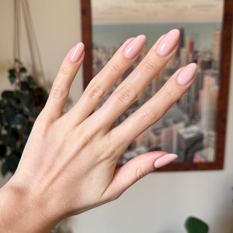 Hand with almond-shaped nails painted in KAG - Gel neutral peachy pink gel polish against blurred cityscape background