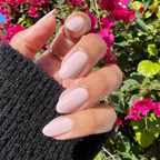 Hand with medium almond nails polished in Kitten for a shiny, neutral-toned manicure outdoors with bright pink flowers in background