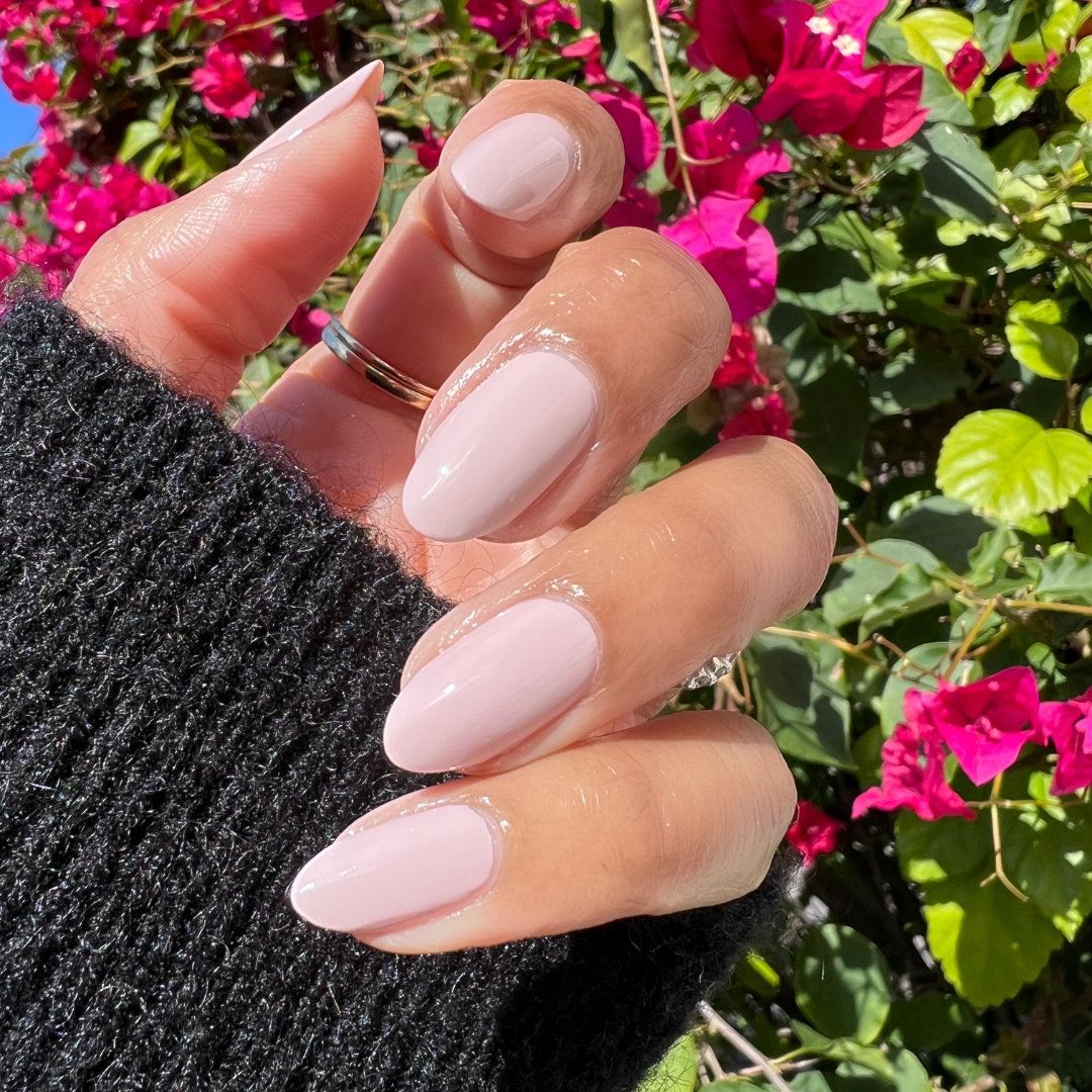 Hand with medium almond nails polished in Kitten for a shiny, neutral-toned manicure outdoors with bright pink flowers in background